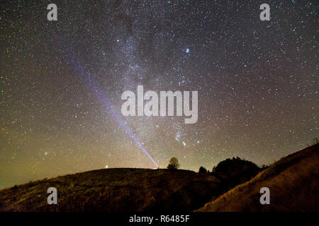 Small silhouette of man with flashlight on dark green grassy hill under beautiful dark blue summer starry sky. Night photography, beauty of nature con Stock Photo