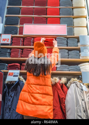 The girl holds out her hand for clothes on the store shelf. shopper pulls on a thing in a clothing store. Rear view of woman who holds out a hand to a shelf with clothes displayed in store. Stock Photo