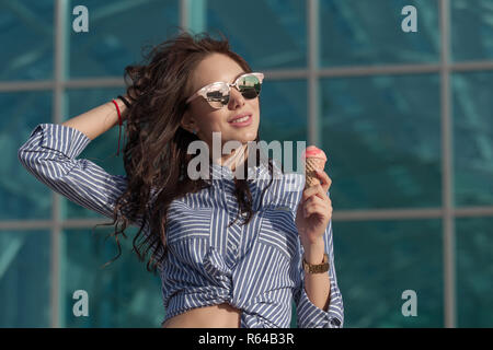 Young, beautiful brunette woman eating pink ice cream in a waffle horn. She's in a good mood She wears glasses with dark glasses, and a shirt in a blue and white strip. Close-up photo Stock Photo