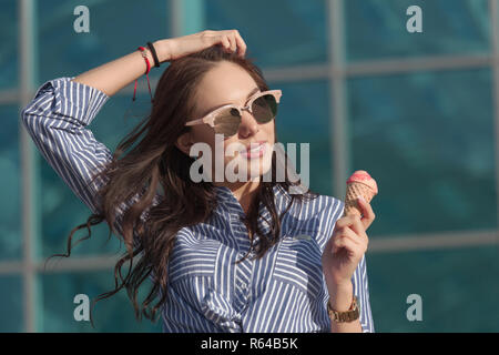 Young, beautiful brunette woman eating pink ice cream in a waffle horn. She's in a good mood She wears glasses with dark glasses, and a shirt in a blue and white strip. Close-up photo Stock Photo