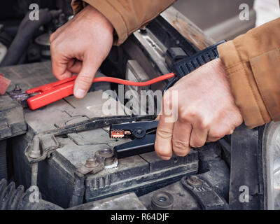 Man recharging a dead car battery in winter time using start-charger. Close up hands of a man with two contact clips from battery charger for vehicles. People under bonnet with starter cables Stock Photo