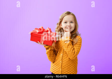 Portrait of a little girl in a warm sweater on a proton purple background. The child looks dreamily up and holds a red box with a gift. The concept of Stock Photo