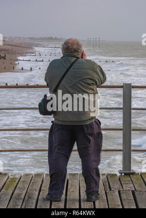 an older man taking photographs of the seaside and rough weather on Bognor regis pier and beach in a storm. Stock Photo