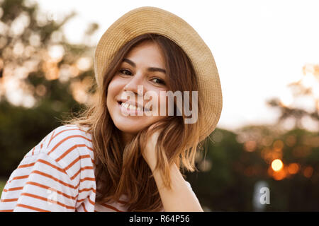 Close up of lovely young girl in summer hat sitting on grass at the park, looking at camera Stock Photo