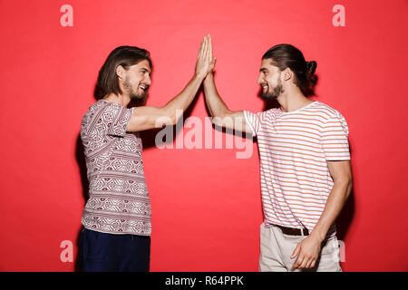 Portrait of two smiling twin brothers standing isolated over red background, giving high five Stock Photo