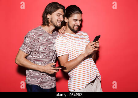 Portrait of a two young smiling twin brothers isolated over red background, using mobile phone Stock Photo