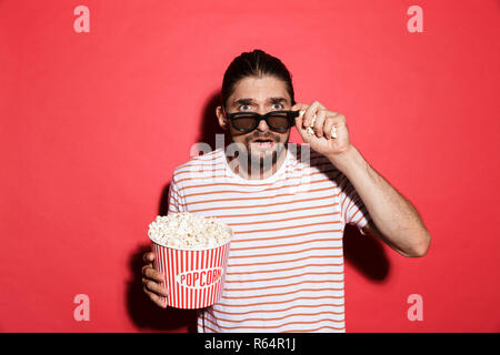 Portrait of shocked man in 3d eyeglasses watching movie, eating popcorn isolated over red background Stock Photo