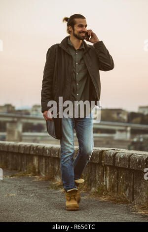 Handsome modern businessman with laptop bag for laptop talking on the phone and walking by the river. Stock Photo