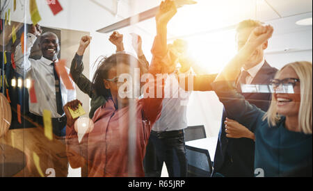 Diverse group of businesspeople cheering and celebrating a winning idea together while brainstorming with sticky notes on a glass wall in a modern off Stock Photo