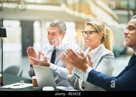 Smiling group of mature businesspeople sitting together at a table in a modern office clapping hands during a seminar Stock Photo