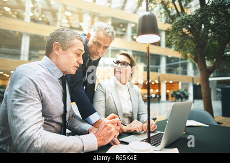 Mature businesspeople having a meeting together in the lobby of a modern office discussing work over a laptop Stock Photo