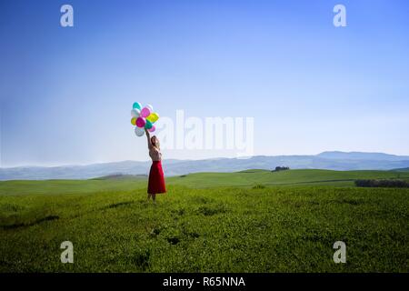 Happy girl in the meadows tuscan with colorful balloons, against the blue sky and green meadow. Tuscany, Italy Stock Photo