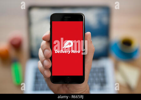 A man looks at his iPhone which displays the Delivery Hero logo, while sat at his computer desk (Editorial use only). Stock Photo