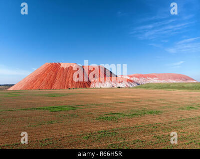 Summer landscape. Red mountain, blue sky, green field. Slag heap as mountains. Big piles of potash waste. Extraction mining potassium salt. Industrial Stock Photo
