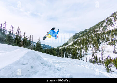 Snowboarder freerider jumping from a snow ramp in the sun on a background of forest and mountains Stock Photo