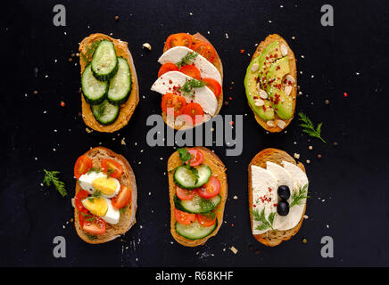 Set of fresh sandwiches snacks on dark bread with cheese, tomatoes, cucumber, boiled eggs and avocado, viewed from above on black background, decorate Stock Photo