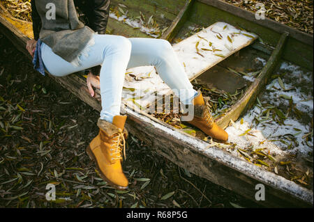 slim girl's legs in blue jeans and brown boots. girl sat on the upturned boat, on the shore, yellow autumn leaves Stock Photo