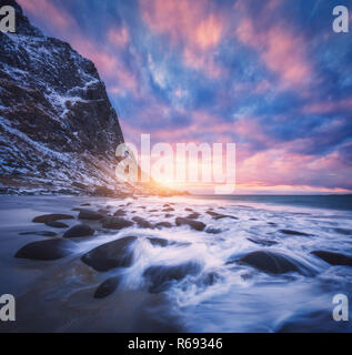 Sandy beach with stones in blurred water, blue sky with colorful clouds and snowy mountains at sunset. Utakleiv beach, Lofoten islands, Norway. Winter Stock Photo