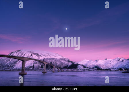 Beautiful bridge against snowy mountains, purple sky with pink clouds and moon at night in Lofoten islands, Norway. Winter landscape with road, blue s Stock Photo