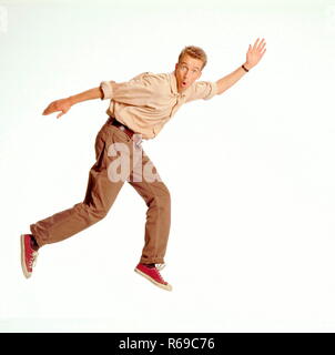 Portrait, Ganzfigur, junger Mann mit kurzen blonden Haaren bekleidet mit brauner Jeans, Hemd und Turnschuhen springt in die Luft Stock Photo