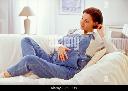 Portrait, hochschwangere Frau sitzt entspannt auf ihrem Sofa und beruehrt ihren Bauch Stock Photo