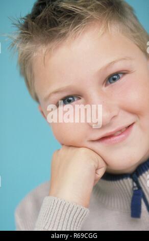 Portrait, Nahaufnahme, grinsender dunkelblonder Junge mit blauen Augen, 9 Jahre, stuetzt sein Kinn auf seine Hand Stock Photo