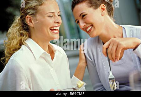 Portrait, 2 junge Fraue, blond und bruenett, sitzen lachend bei Kaese mit Gabeln in den Haenden in einer Bar Stock Photo