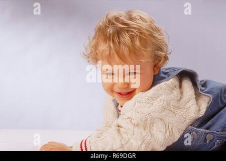 Portrait,  Innenraum, krabbelnder 1-jaehriger blonder Junge bekleidet mit Jeanslatzhose und weissem Pullover Stock Photo