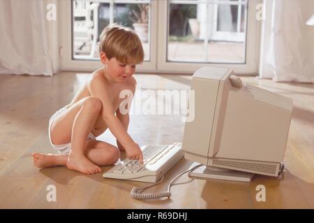 Portrait,  Innenraum, 6-jaehriger blonder Junge bekleidet nur mit Unterhose sitzt auf dem Holzboden und spielt am Computer Stock Photo