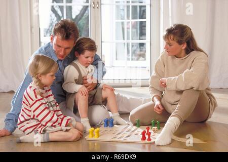 Innenraum, Ganzfigur, Familie mit 2 Kindern sitzt auf dem Fussboden vor dem Fenster und spielt eine Partie Mensch-aergere-dich-nicht Stock Photo