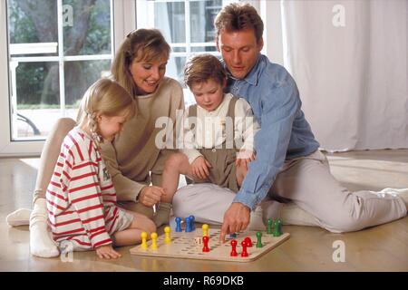Innenraum, Ganzfigur, Familie mit 2 Kindern sitzt auf dem Fussboden vor dem Fenster und spielt eine Partie Mensch-aergere-dich-nicht Stock Photo