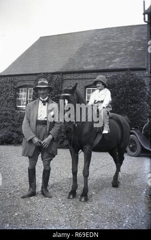 1930s, historical, on a gravel driveway outside a country house, an elderly man in riding gear standing next to a little girl on a pony or small horse, also wearing riding gear, probably her grandfather helping her learn to ride. Stock Photo