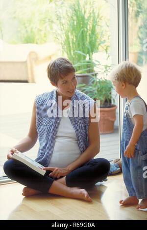 Portrait, Ganzfigur, schwangere Frau mit kurzen braunen Haaren sitzt mit einem Buch in der Hand vor der offenen Tuer auf dem Boden und spricht mit ihrem 2-jaehrigen Sohn Stock Photo