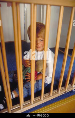 Portrait,  Innenraum, 2-jaehriger blonder Junge bekleidet mit Jeans und weissem Pullover sitzt mit seinem Teddy im Arm hinter einem Treppengitter Stock Photo