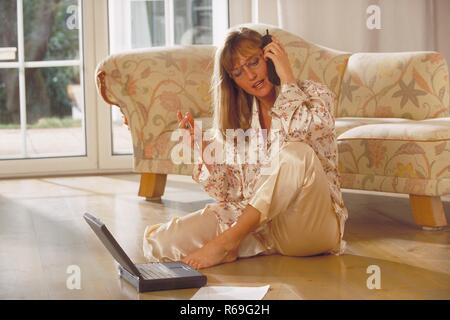 Innenraum, Portrait, Ganzfigur, Frau Mitte 30 mit langen blonden Haaren und Brille sitzt im Schlafanzug mit ihrem Laptop vor dem Sofa auf dem Fussboden und telefoniert Stock Photo