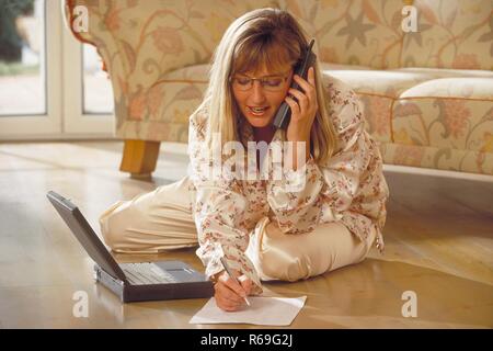 Innenraum, Portrait, Ganzfigur, Frau Mitte 30 mit langen blonden Haaren und Brille sitzt im Schlafanzug mit ihrem Laptop vor dem Sofa auf dem Fussboden, telefoniert und macht sich Notizen Stock Photo