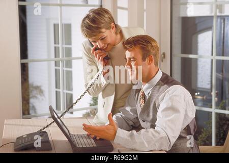Innenraum, blondes Paar Mitte 30, der Mann sitzt am Tisch in der Naehe des Fensters am Laptop, waehrend die Frau mit telefoniert Stock Photo
