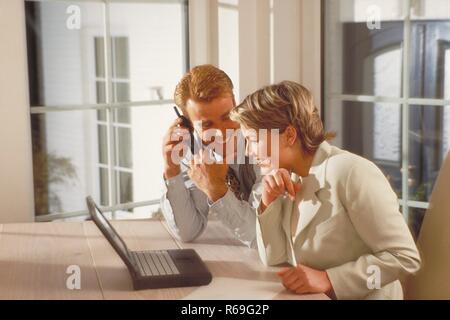 Innenraum, blondes Paar Mitte 30 sitzt lachend am Tisch in der Naehe des Fensters vor dem Laptop, waehrend der Mann mit einem Handy telefoniert Stock Photo