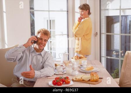 Innenraum, Halbfigur, Paar Mitte 30, Mann sitzt am gedeckten Fruehstueckstisch und telefoniert mit einem Handy, sie steht am Fenster und beobachtet ihn skeptisch Stock Photo