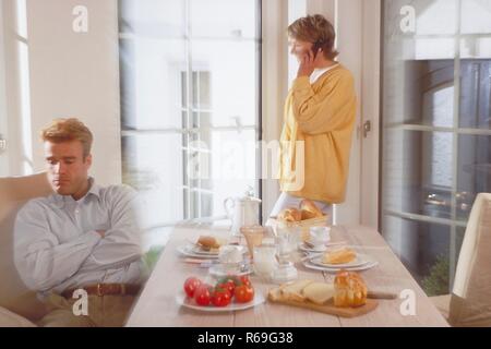 Innenraum, Halbfigur, Paar Mitte 30, Mann sitzt genervt am gedeckten Fruehstueckstisch, sie steht am Fenster und telefoniert mit einem Handy Stock Photo