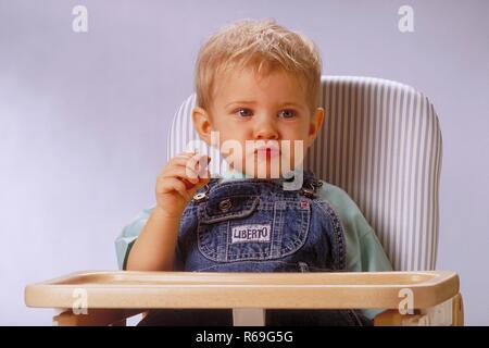 Portrait,  Innenraum, ca. 1-jaehriger blonder Junge bekleidet mit Jeanslatzhose sitzt mit einem Wuerstchen in der Hand in einem Hochstuhl Stock Photo