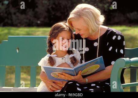 Outdoor, blonde Frau Mitte 60 bekleidet mit schwarzen Kleid mit weissen Punkten und ihre 6 Jahre alte  Enkeltochter sitzen im Garten auf einer blaugruenen Bank  und lesen ein Buch Stock Photo