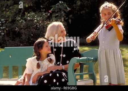 Outdoor, blondes 10 Jahre altes Maedchen spielt ihrer Oma und ihrer kleinen  Schwester, die im Garten auf einer Bank sitzen, eine Melodie auf der Geige vor Stock Photo