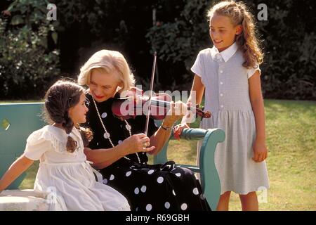 Outdoor, blonde Frau Mitte 60 bekleidet mit schwarzen Kleid mit weissen Punkten sitzt mit  ihrer 2  Enkeltoechtern im Garten auf einer blaugruenen Bank und spielt eine Melodie auf einer Geige Stock Photo