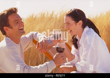 Portrait, Halbfigur, junge Frau mit Sommersprossen und langen rotbraunen Haaren, traegt Sonnenbrille und weisses Hemd, sitzt mit ihrem Partner entspannt mit einem Glas Rotwein auf einem hoelzernen Liegestuhl zwischen Graesern in den Duenen Stock Photo