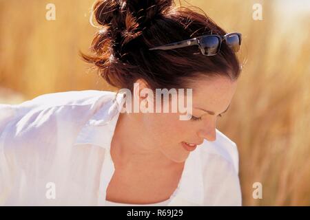 Portrait, Profil einer jungen Frau mit Sommersprossen und hochgesteckten rotbraunen Haaren und Sonnenbrille auf der Stirn, bekleidet mit weissem Hemd, sitzt entspannt zwischen goldfarbenen Graesern in den Duenen