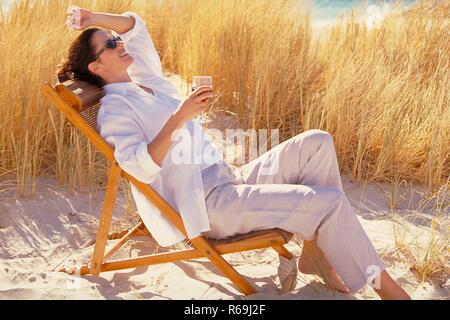 Portrait, Vollfigur, junge Frau mit Sommersprossen und hochgesteckten rotbraunen Haaren, traegt Sonnenbrille, weisses Hemd und helle Hose, sitzt entspannt mit einem Glas Rotwein auf einem hoelzernen Liegestuhl zwischen Graesern in den Duenen Stock Photo