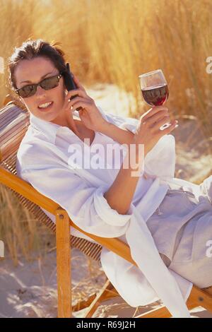 Portrait, Halblfigur, junge Frau mit Sommersprossen und hochgesteckten rotbraunen Haaren, traegt Sonnenbrille, weisses Hemd und helle Hose, sitzt entspannt mit einem Glas Rotwein auf einem Liegestuhl zwischen Graesern in den Duenen und telefoniert Stock Photo