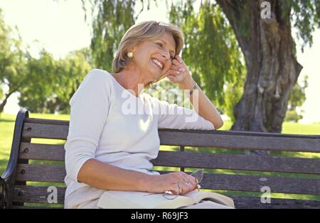 Portrait, Parkszene, blonde Frau, Mitte 50, bekleidet mit heller Hose und Top, sitzt entspannt mit einem Buch und ihrer Lesebrille in der Hand auf einer Parkbank unter einem Baum