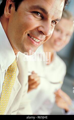 Strassenszene, Portrait eines laechelnden bruenetten jungen Mannes, ca. 30 Jahre, bekleidet mit weissem Hemd und goldfarbener Krawatte, im Hintergrund eine hell gekleidete Frau mit blonden Locken Stock Photo